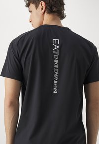 Čierne tričko s krátkymi rukávmi, ktoré má vzadu vertikálny biely potlač loga. Hladká tkanina, uvoľnený strih a kruhový výstrih.