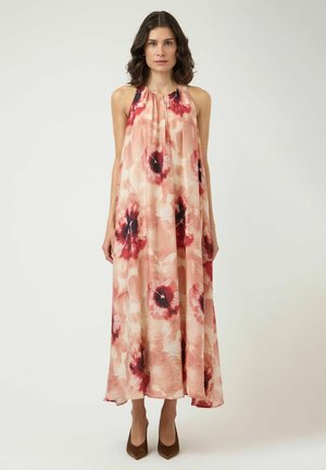 Kobieta ubrana w bezrękawową, luźną, kwiecistą sukienkę maxi w różowych, czerwonych i beżowych tonacjach, z brązowymi, spiczastymi obcasami, stojąca na jednolitym tle.