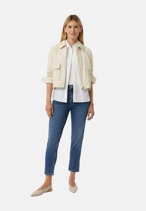 Vrouw staat in een beige cropped jacket over een wit overhemd, blauwe cropped jeans en beige slingback flats.
