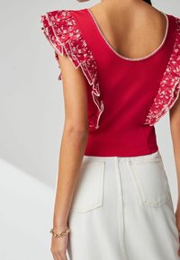 Haut rouge côtelé avec des manches à volants floraux brodés, présentant un dos à décolleté arrondi. Associé à un jean taille haute de couleur claire.