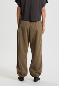 Vapaasti istuvat, oliivinvihreät housut, joissa on joustavat hemmet nilkoissa. Kangas vaikuttaa sileältä, mutta siinä on hienovarainen tekstuuri. Yhdistetty lyhyen, tumman yläosan kanssa.