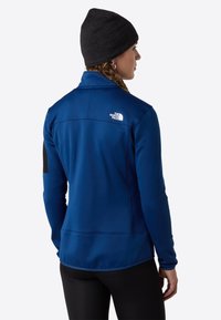 Modrá fleecová bunda s vysokým límcem, kapsou na zip a černým akcentem na rukávu. Měkká textura, lehký design.
