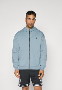 Lys blå zip-up hoodie lavet af blødt stof, med en kængurulomme og et sort logo på brystet. Båret med sorte mesh-shorts.