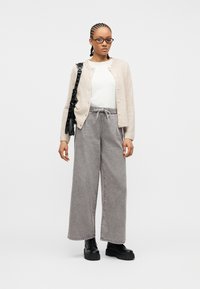 Γυναίκα με πλεξούδες κόρνροου, φορά γυαλιά, μπεζ ζακέτα, λευκό τοπ, γκρι παντελόνι φαρδιάς γραμμής, μαύρες μπότες και τσάντα ώμου, στέκεται μπροστά σε λευκό φόντο.