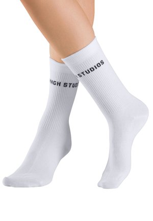 Legs crossed wearing white ribbed crew socks with black text reading "STUDIOS" near the top against a white background.