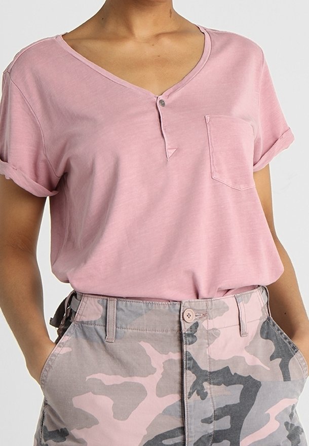 Femme portant un t-shirt rose à manches retroussées, col en V avec poche avant et un pantalon camouflage rose et gris taille haute, les mains dans les poches.