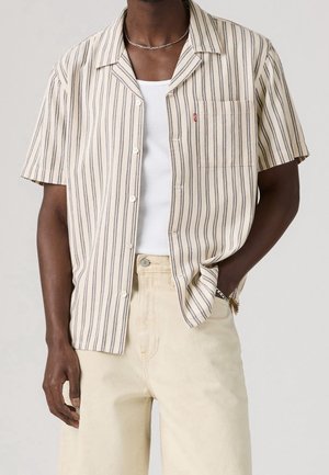 Mann trägt beige und schwarz vertikal gestreiftes kurzärmeliges Hemd mit Knöpfen, weißes Tanktop, hellbeige Hose, silberne Kettenkette und Armband.