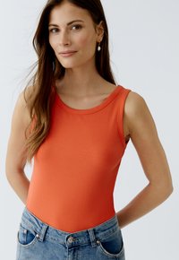 Geribbelde oranje tanktop met een brede ronde hals, gecombineerd met hoge blauwe denim jeans. Het model heeft lang haar en draagt parel-druppelaar oorbellen.