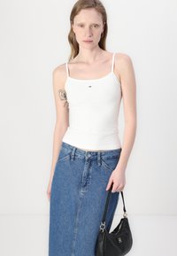 White fitted camisole with thin straps, featuring a small logo detail on the chest, paired with a mid-length blue denim skirt and a black handbag.