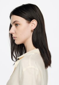 Aretes de aro dorados en una persona con cabello largo y oscuro, vistiendo una camisa ligera de color beige claro. El fondo es blanco.