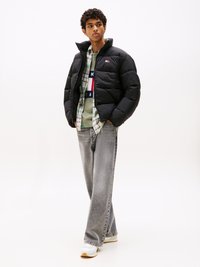 Black puffer jacket with a high collar, paired with a green sweater and plaid shirt. Light gray jeans and white sneakers complete the outfit.