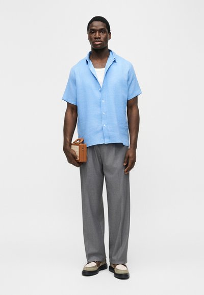 Άνδρας που στέκεται, φοράει ανοιχτό μπλε πουκάμισο με κοντά μανίκια, γκρι παντελόνι, δίχρωμα μοκασίνια, κρατώντας ένα μικρό, καφέ ραδιόφωνο σε vintage στυλ.