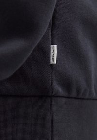Tejido de algodón azul marino con superficie texturizada; presenta una etiqueta blanca con "JACK & JONES" en la costura, destacando la marca.