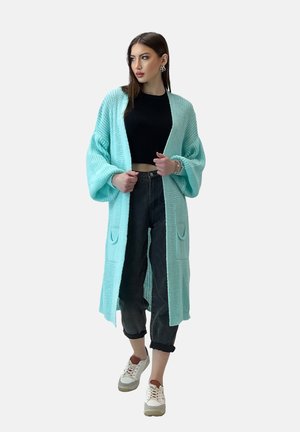 Cardigan lavorato a maglia verde menta con maniche lunghe, tasche frontali e una vestibilità ampia, abbinato a un top corto nero e jeans con risvolto.
