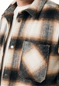 Randig flanellskjorta i kräm, brunt och svart. Har en bröstficka, metalltryckknappar och ett texturerat tyg.