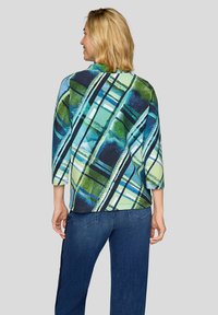 Blonde Frau, die wegschaut, trägt Oberteil mit abstraktem blau-grün-weiß kariertem Muster, dreiviertellangen Ärmeln und dunkelblaue Jeans.