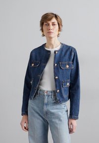 Denim jakna tamno plave boje s okruglim izrezom, zlatnim dugmadi, dva prednja džepa i skraćenim krojem, u kombinaciji sa svijetloplavim farmerkama.