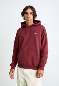 Hoodie bordeaux en coton doux, avec une poche kangourou à l'avant et une capuche avec cordon de serrage. Petite étiquette avec logo sur la poitrine. Manches ajustées.
