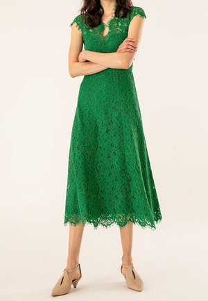 Femme portant une robe midi en dentelle verte avec des manches ballon, debout, bras croisés, assortie à des chaussures beige à talon bas.