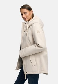 Beige huppari, jossa on pehmeä tekstuuri, vetoketjukiinnitys ja nepparit. Varustettu sivutaskuilla ja merkkipatchilla hihassa.