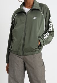 Grön polyester zip-jacka med vita detaljer, med "adidas" logotyp på vänster bröst och ned längs höger ärm. Elastisk midja.