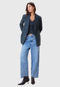Blazer in suede navy sopra un gilet nero, abbinato a jeans baggy azzurri e stivaletti neri alla caviglia. Presenta dettagli con bottoni e orli grezzi.