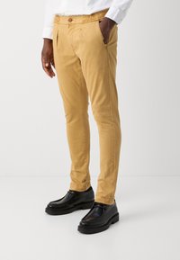 Pantalons beige ajustés en coton avec poches latérales, taille élastique et détail bouton, associés à des chaussures en cuir noires.