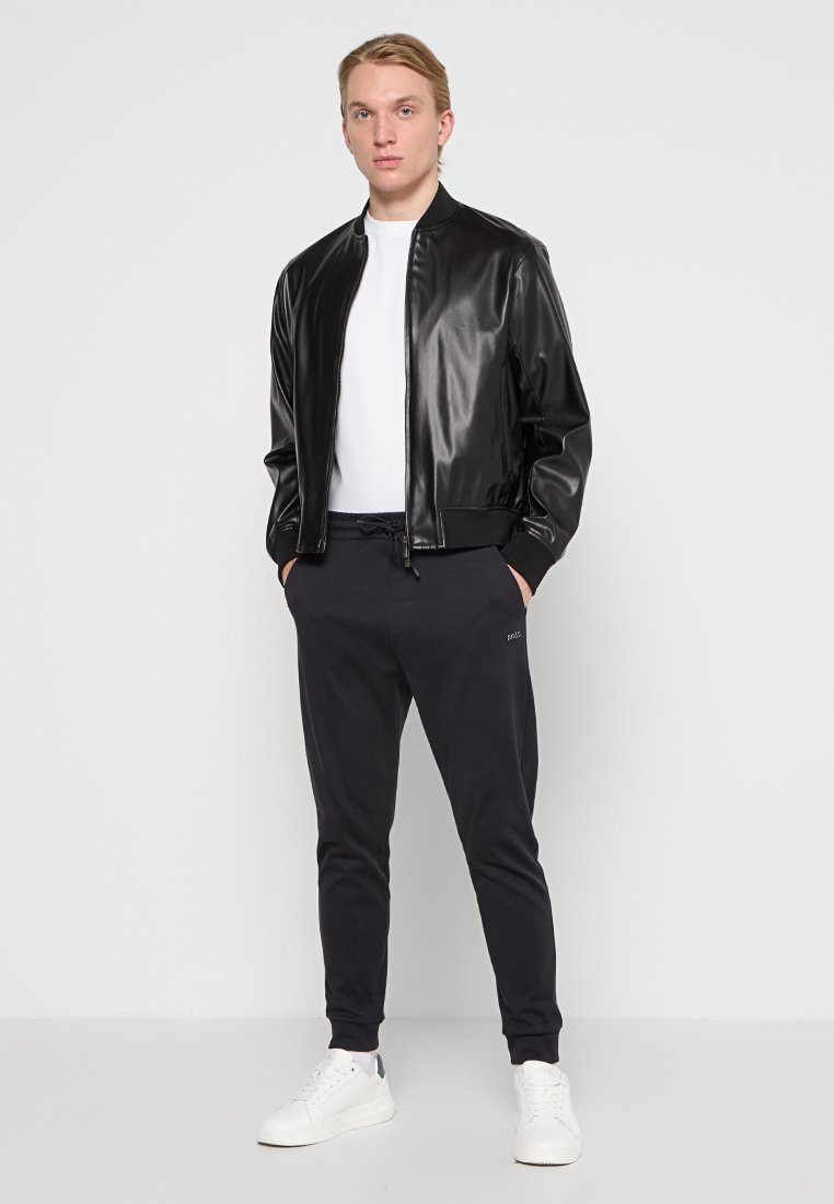 Giubbotto bomber nero e lucido con colletto a coste, abbinato a pantaloni jogger scuri e sneaker bianche. Design pulito, dettagli minimi visibili.