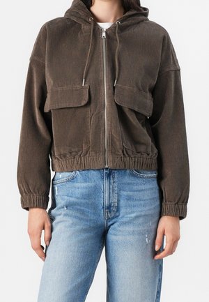 Osoba ubrana w brązową kurtkę z kapturem zapinaną na suwak, wykonaną z sztruksu, z kieszeniami na klapki oraz niebieskie jeansy z efektem sprania.