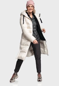 Lange witte pufferjas met een capuchon, gecombineerd met een zwart-wit gestreept shirt, donkere broeken en stijlvolle, meerkleurige sneakers.