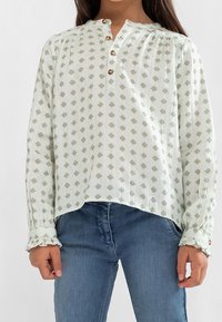 Light green blouse with small floral pattern, round neckline, button placket, and ruffled cuffs; made from a textured fabric.
