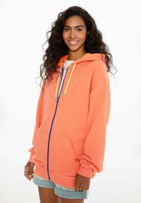 Mikina s kapucňou vyrobená z jasne oranžovej textílie s modrým zipsom a žltými šnúrkami, s predným vreckom a voľným strihom.