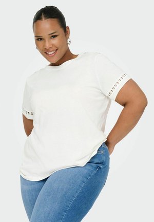 Camiseta de algodón blanca de mangas cortas, con detalles de encaje en los dobladillos de las mangas. Combinada con unos jeans de mezclilla azules.