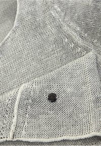 Tissu gris clair tricoté avec des coutures visibles et une petite étiquette ronde noire cousue dessus.