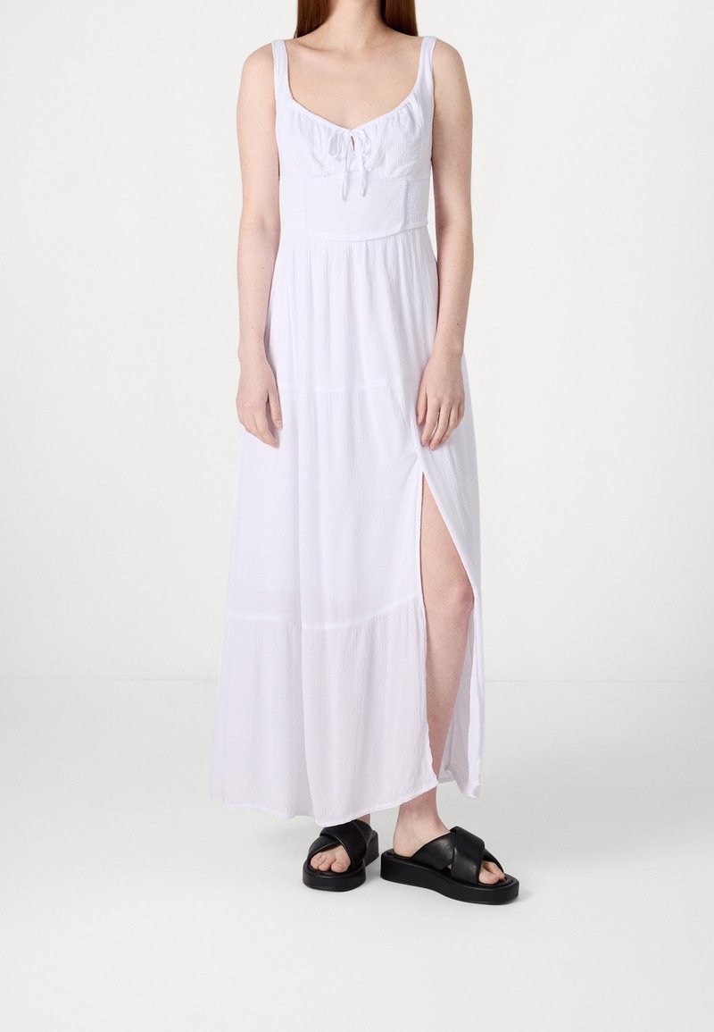 Vit maxiklänning med rundad halsringning, draperad byst och hög slits till låret. Lättviktsmaterial, ärmlös design och avslappnad passform. Svarta platåsandaler.
