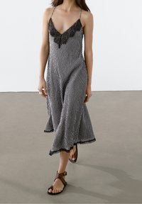 Vrouw in een zwart-wit geruit slipdress met kant en bruine sleehak sandalen, wandelend over een gladde grijze vloer.