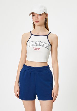 Hvid cropped tanktop med marineblå kant og rød print, parret med blå shorts med elastisk talje og sidelommer. Hvid kasket, hoops øreringe.
