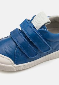 Adidași din piele albastră cu două benzi Velcro, talpă din cauciuc alb, laterale perforate și o etichetă gri pe limbă. Include cusături contrastante.
