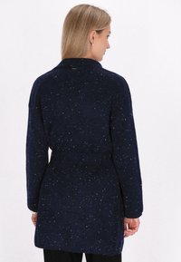 Marinblå, långärmad cardigan med en texturerad yta. Har en bältad midja och en klassisk krage. Spräcklig detalj ger visuell intress.