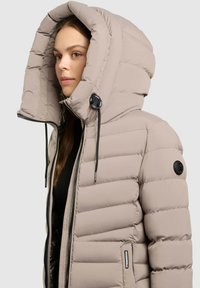 Een lichtbeige pufferjack met een gewatteerd ontwerp, een oversized capuchon, ritssluiting en strikkoorden. Bevat een logo-opdruk op de linkermouw.