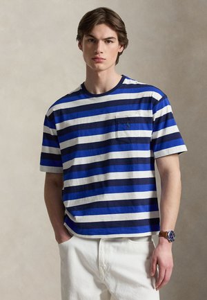 Jeune homme portant une chemise à manches courtes rayée bleu, marine et blanc avec une poche poitrine et un pantalon blanc, debout devant un fond neutre.