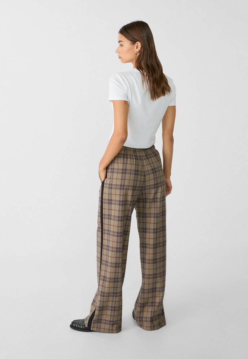 Donna con lunghi capelli castani, che indossa una maglietta bianca aderente a maniche corte e pantaloni largo a quadri marroni con striscia nera e scarpe nere.