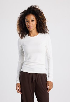 Top blanco de manga larga con un diseño ajustado, textura acanalada y cuello redondo, combinado con pantalones marrones de corte relajado. Detalle de marca minimalista.