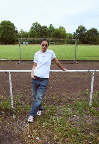White t-shirt with blue logo, relaxed-fit blue jeans, and white/red athletic shoes. Standing by a chain-link fence on a grassy field.