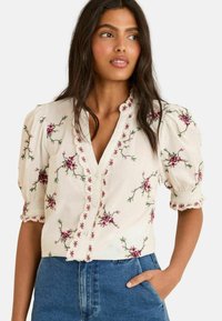 White blouse with short puffed sleeves, featuring pink floral embroidery and scalloped button placket. Paired with denim jeans.