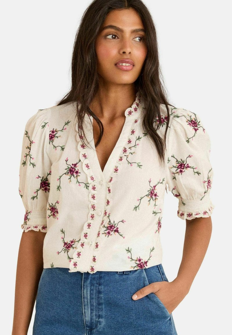 White blouse with short puffed sleeves, featuring pink floral embroidery and scalloped button placket. Paired with denim jeans.