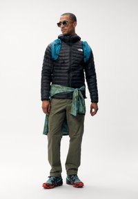 Zwarte pufferjack, teal rugzakken, olijfgroene cargobroeken, gestreept blauw shirt geknoopt om de taille, en meerkleurige trailrunschoenen.