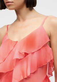 Γυναίκα που φοράει ροζ κοραλί αμάνικη μπλούζα με στρωματώδεις βολάν και λεπτές τιράντες σπαγγέτι, που δείχνει τον λαιμό και την περιοχή των ώμων.