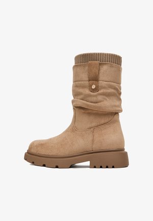 Bota de ante beige hasta mitad de la pantorrilla con puño de punto acanalado, lengüeta para tirar, punta redondeada y suela gruesa de goma con surcos profundos para mayor agarre.