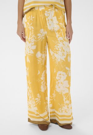 Żółte szerokie spodnie z białym kwiatowym wzorem i paskiem w paski na dole, noszone z brązowymi plecionymi butami, pokazane od talii do stóp.